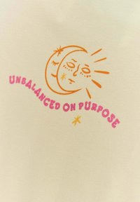 Krémová tkanina s grafickým designem slunce a měsíce s obličejovými rysy, doplněná textem "NEVYVÁŽENÝ ÚMYSLNĚ" v růžové barvě.