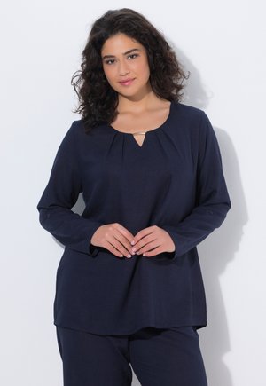 Kvinde med krøllet hår iført en marineblå langærmet top og matchende bukser, stående med hænderne blidt foldet foran mod en hvid baggrund.