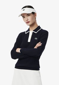 Polo en tricot bleu marine avec des accents crème et une patte de boutonnage ; présente un logo Lacoste vert sur la poitrine gauche. Une visière blanche complète la tenue.