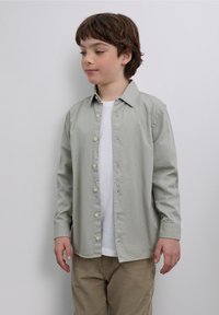Jasnozielona koszula na guziki wykonana z bawełny, z klasycznym kołnierzykiem, długimi rękawami i jasnożółtymi guzikami, noszona na białej koszulce.