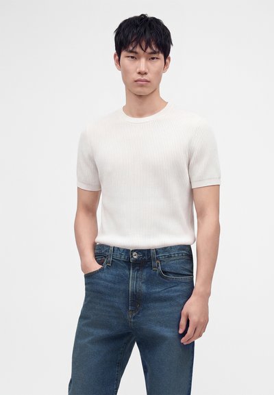 Jeune homme aux cheveux noirs courts portant un t-shirt blanc côtelé à manches courtes et un jean bleu, debout avec une main dans la poche.