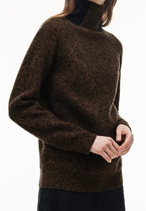 Brązowy, teksturowany sweter z golfem wykonany z wełny, noszony przez osobę z rękami złączonymi z przodu, w połączeniu z ciemnymi spodniami.