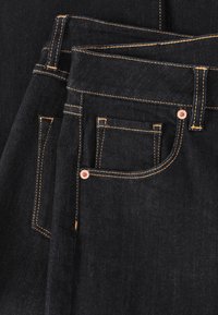 Jeans din denim închis, pliați, cu cusături portocalii, un mic buzunar în față și cu nituri din cupru, având o textură netedă și un design clasic.