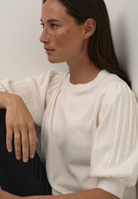 Pull blanc avec des manches bouffantes à côtes. Texture douce, col rond, associé à un pantalon foncé. Design simple et moderne.