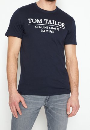 Man draagt een marineblauw T-shirt met de tekst "Tom Tailor Genuine Crafts Est 1962" en lichtgrijze jeans, staand met de armen ontspannen langs het lichaam.