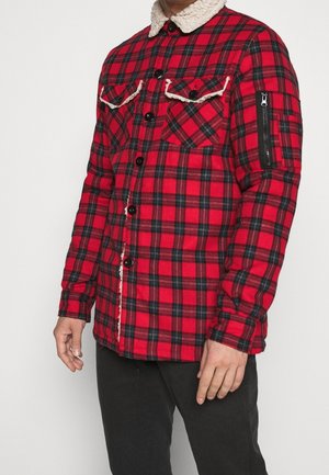 Mężczyzna w czerwonej i czarno kratowanej kurtce zapinanej na guziki z kołnierzem z materiału sherpa i kieszenią na suwak na rękawie, w połączeniu z czarnymi spodniami.