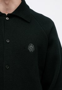 Cardigan negru tricotat cu guler și nasturi, având un mic emblemă brodată cu un gândac pe partea stângă a pieptului.