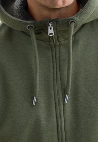 Grüne zip-up Kapuzenjacke aus weichem Material, mit flauschiger Innenauskleidung, Kordelzugkapuze und metallischen Reißverschlussakzenten.