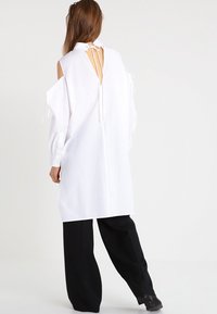 Woman standing with back turned, wearing a white long-sleeve open-back shirt with shoulder cutouts and loose black pants.