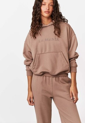 Kobieta w brązowej bluzie z kapturem z napisem "LA MANIA" i dopasowanych spodniach, stojąca na jednolitym białym tle.