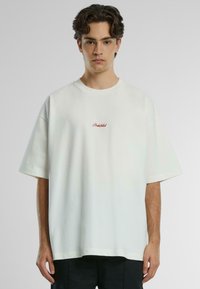 Tricou oversize din bumbac alb, cu mâneci scurte și un mic broderie roșie "Interzis" pe piept. Textură netedă, croială lejeră.