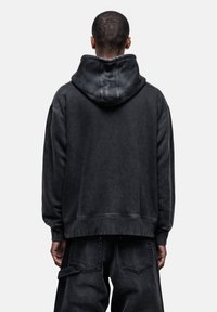 Felpa nera con una vestibilità morbida e un cappuccio ampio, caratterizzata da una texture soffice e dettagli di cucitura visibili. Indossata con jeans scuri abbinati.
