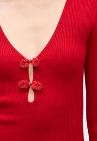 Rød ribbet topp med dyp V-hals, som har to utsmykkede røde dekorasjoner ved halslinjen og en nøkkelhullsåpning.