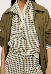 Wieloczęściowy zestaw z oliwkową zieloną kurtką, nałożoną na bluzkę w kratę w białym, brązowym i zielonym kolorze. Teksturowany materiał z guzikami.