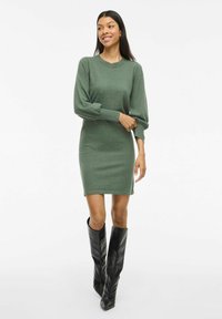 Grünes Strickkleid mit langen, gepufften Ärmeln und figurbetonter Silhouette, kombiniert mit kniehohen schwarzen Stiefeln mit Absatz. Glatte Textur und schlichtes Design.