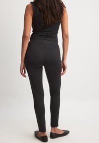 Melna bezpeļu topa kombinācija ar melnām legingām, abiem izgatavotiem no gludas, elastīgas auduma. Funkcijas ietver pieguļošu dizainu un plakanu šuvi, valkājot ar melnu plakanajā zābakos.