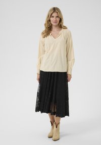 Blouse crème clair avec un col en V et des manches froncées, associée à une longue jupe en dentelle noire. Chaussures : bottes beiges.