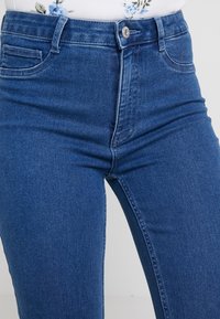 Jeans de mezclilla azul de cintura alta con cinco bolsillos, cierre frontal de botón y un acabado suave con ligera textura.
