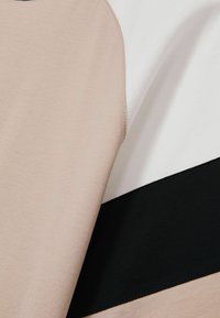 Krāsu bloku tops, kas sastāv no bēšiem, baltiem un melniem segmentiem, izgatavots no mīkstas auduma ar gludu tekstūru un kontrastējošām šuvēm.