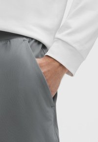 Graue Sportshorts mit einer glatten Textur, die mit einer Seitentasche ausgestattet sind. Ein weißes Langarmshirt mit gerippten Bündchen ist teilweise sichtbar.