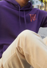 Fialová mikina s přední kapsou, na které se nachází červené logo. Kombinováno se světlými texturovanými kalhotami. Pozadí zobrazuje venkovní prostředí.