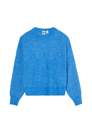 Vrouw met een lavendelkleurige gebreide muts en een felblauwe trui, houdt een meerkleurige deken vast, met haar rug naar een witte achtergrond gekeerd.