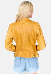 Žlutá kožená bunda s přiléhavým střihem. Má ramenní vycpávky, nastavitelné přezky v pase a hladký, texturovaný povrch.
