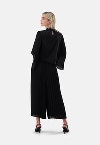 Schwarzer, plissierter Jumpsuit mit langen, transparenten Ärmeln, einem Schlüsselloch-Ausschnitt am Rücken und weiten Hosenbeinen, kombiniert mit Riemchen-Hochabsatzschuhen.