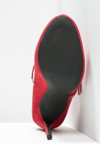 Red high-heeled shoe with a smooth suede texture and a sleek pointed toe. Black rubber sole with a textured grip. Minimalist design.