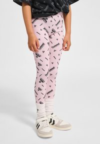 Różowe legginsy z czarnym, powtarzającym się wzorem logo, w połączeniu z białymi skarpetkami i kremowymi sneakersami z czarnymi paskami.