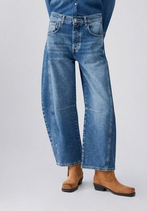 Persona che indossa jeans a gamba larga blu e stivaletti in camoscio color cuoio su uno sfondo grigio chiaro semplice.