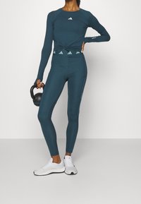Teal långärmad träningstopp med knuten front, i kombination med högmidjade teal leggings. Vita träningsskor och en svart kettlebell.