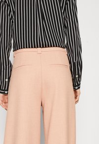 Růžové široké kalhoty s hladkou texturou, s čistým pasem a dvěma zadními kapsami, nošené s černobílým pruhovaným tričkem.