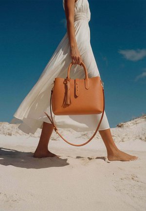 Femme en robe blanche fluide marchant pieds nus sur le sable, tenant un grand sac à main en cuir marron clair avec un détail de gland, devant un ciel bleu clair.