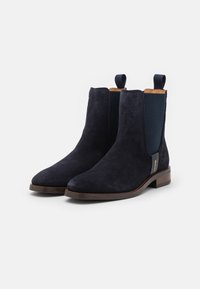 Botas Chelsea de ante azul marino con paneles laterales elásticos, tiradores, textura suave y suela de cuero marrón con tacón bajo.