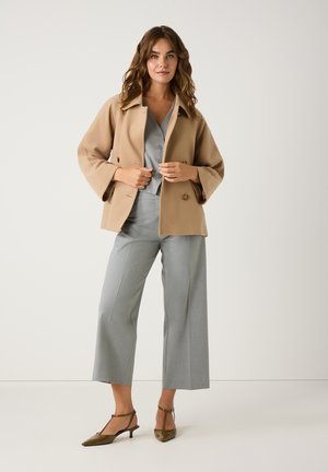 Beige dubbelzijdig jasje met brede mouwen, gecombineerd met een grijze blouse met knopen en bijpassende wijduitlopende broek. Bruine puntige hakken.