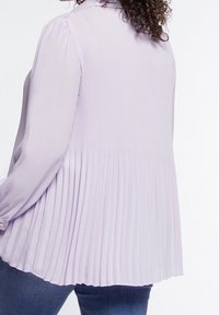 Ljuslila långärmad blus med plisserad baksida, slät textur, knappar vid manschetterna och en avslappnad passform. Lämplig för vardagsbruk.
