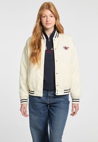 Witte bomberjack met marineblauwe strepen op de manchet en onderkant. Voorzien van een drukknoopsluiting en geborduurd logo detail. Draagt over een zwarte top.