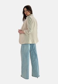 Blazer strutturato beige con maniche arrotolate e due tasche frontali, abbinato a pantaloni in denim a gamba larga di colore azzurro chiaro con orlo sfilacciato.