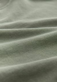 Gros plan sur un tissu lisse vert clair avec des plis doux et une texture fine de tricot visible, mettant en valeur la qualité du matériau et le détail du tissage.