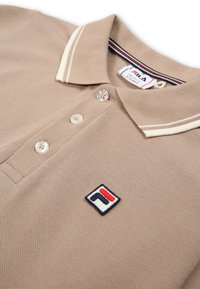 Beige polo-paita, jossa on teksturoitu kangas, kahden napin kaulus, kontrastiväriset raidalliset kaulukset ja brodeerattu logo vasemmassa rintapielessä.
