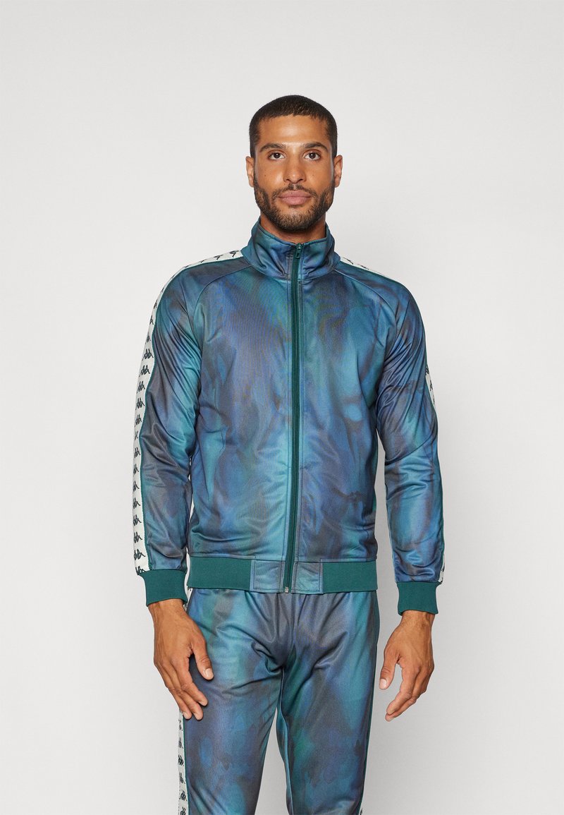 Chándal para hombre con cremallera en un patrón degradado azul-verde, fabricado en material sintético suave, con franjas laterales en contraste y puños acanalados.