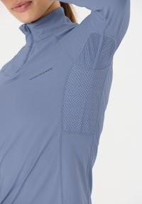 Himmelblaues sportliches Zip-Up-Oberteil mit einem strukturierten Mesh-Einsatz unter dem Arm, versehen mit dezenten Nähten und dem Wort "ENDURANCE", das auf der Brust gedruckt ist.