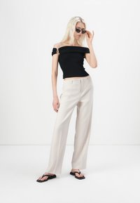 Czarna bluzka typu off-shoulder z prążkowanej tkaniny w połączeniu z jasnobeżowymi spodniami wide-leg oraz czarnymi klapkami, na neutralnym tle.