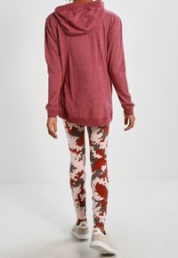 Red hoodie with a low hem and deep hood, paired with pink leggings featuring a red and brown abstract pattern. White athletic shoes.