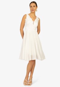 Weißes Kleid mit V-Ausschnitt und floralem Stickerei auf dem Oberteil. Fließender, plissierter Rock in Knielänge, kombiniert mit weißen Riemchensandalen.