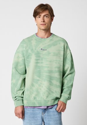 Jeune homme portant un sweat-shirt tie-dye vert clair avec le logo "Faguo", une chemise violette claire en dessous et un jean bleu clair, posant devant un fond blanc.