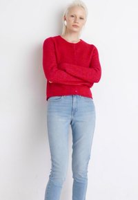 Cardigã vermelho, texturizado, com decote redondo e fecho de botões, combinado com jeans justos azul claro. Modelo de pé com os braços cruzados contra um fundo branco.