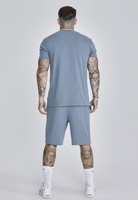 Set de tricou din bumbac albastru și șort. Șorturile au un elastic la talie. Tatuaje notabile vizibile pe brațe și picioare. Pantofi sport albi și șosete purtate.