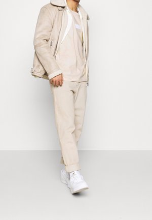 Beige mockajacka med fårskinnskrage, ljusbeige t-shirt och matchande byxor. Vita sneakers kompletterar outfiten.